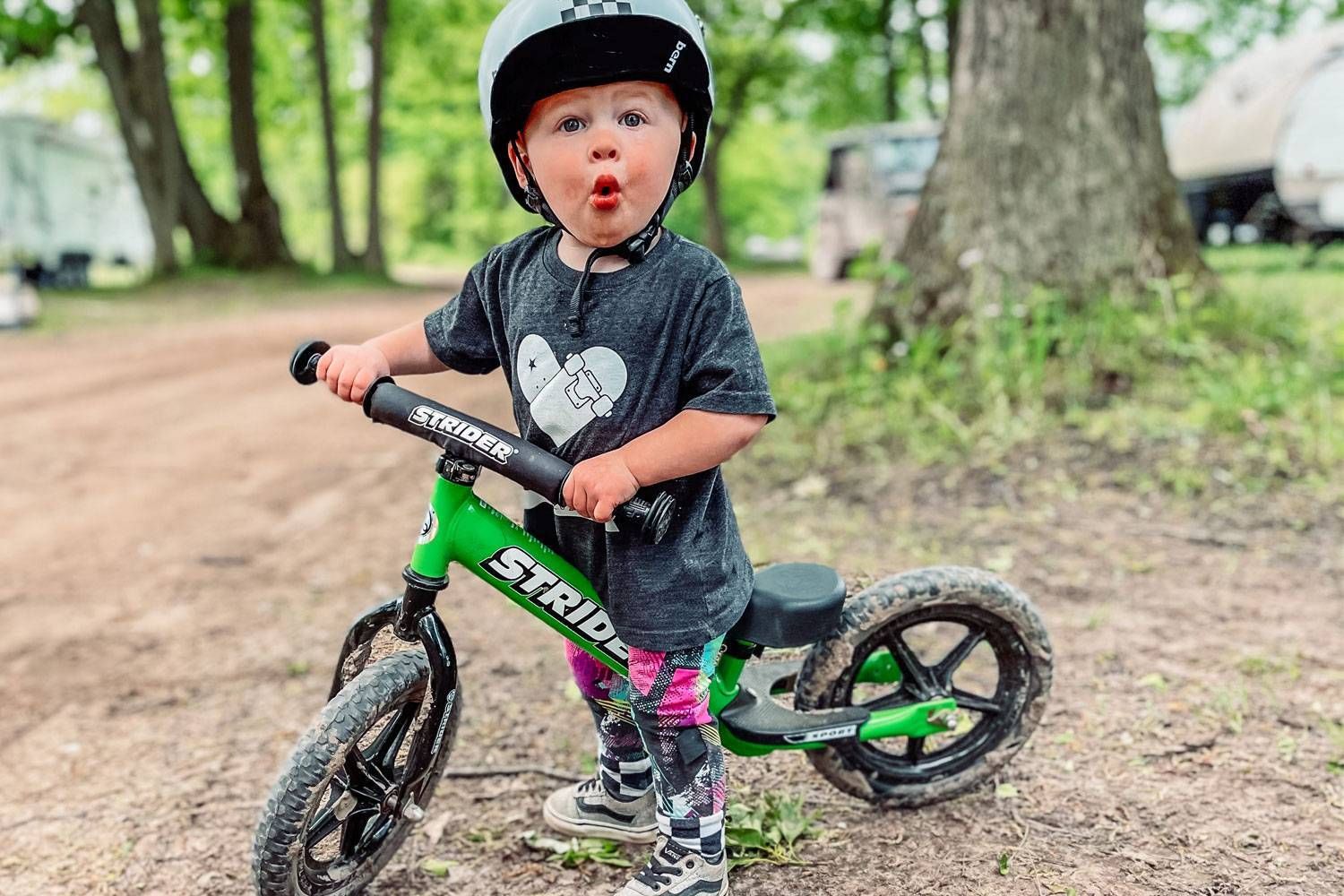 A toddler takes a break from riding a green Strider balance bike