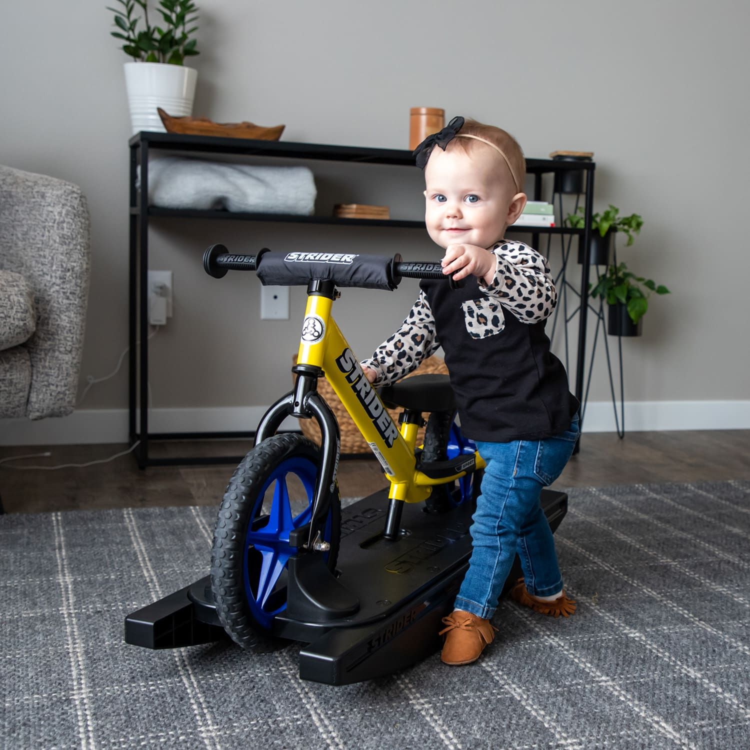 A toddler smiles as she prepares to get on a yellow Strider 12 Sport 2-in-1 Rocking Bike with blue Ultralight Wheels