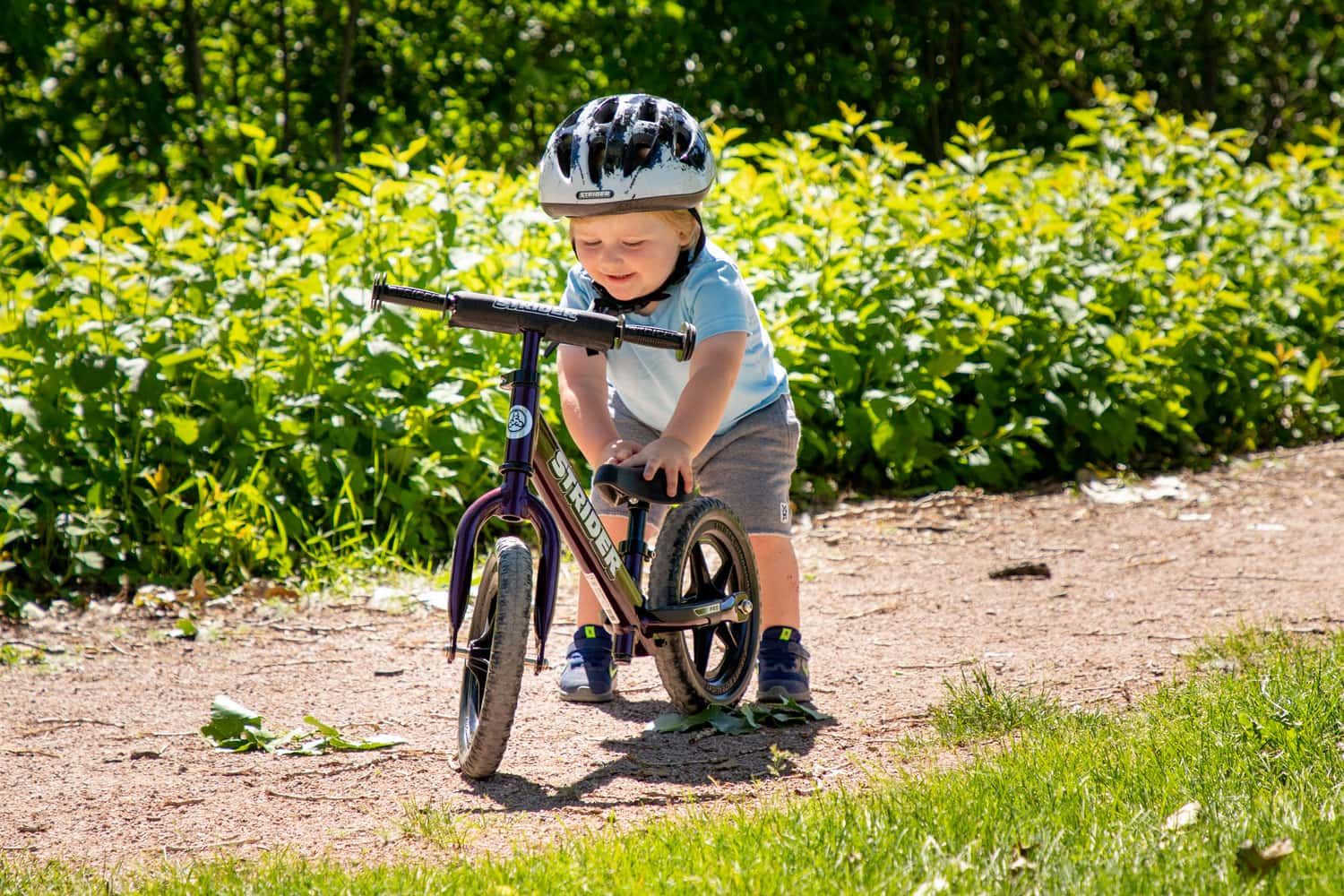 A boy adjusts the seat of a purple Strider 12 Pro balance bike on a gravel trail
