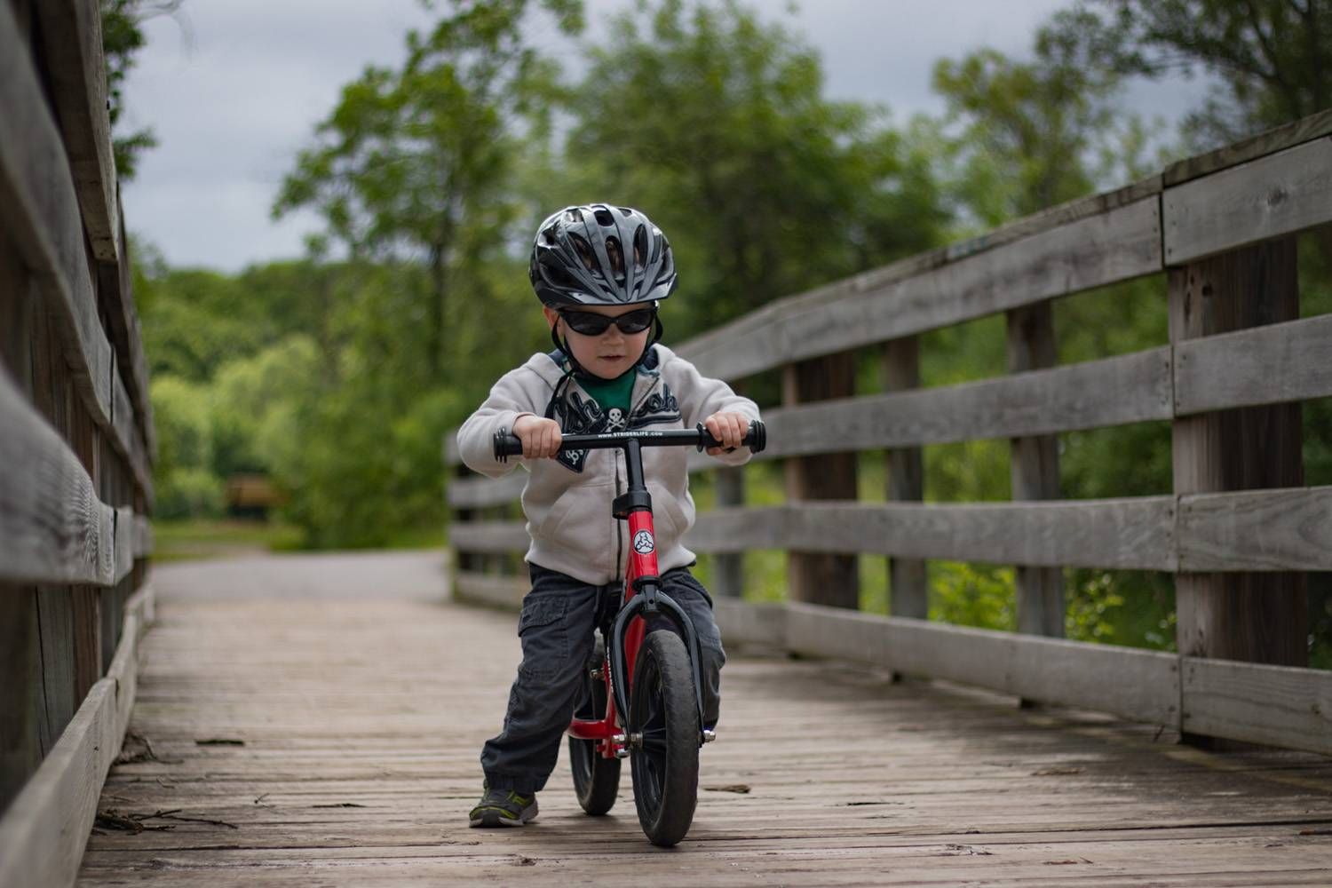 a boy in a black bike helmet riding a red Strider 12 Classic balance bike across a wooden bridge