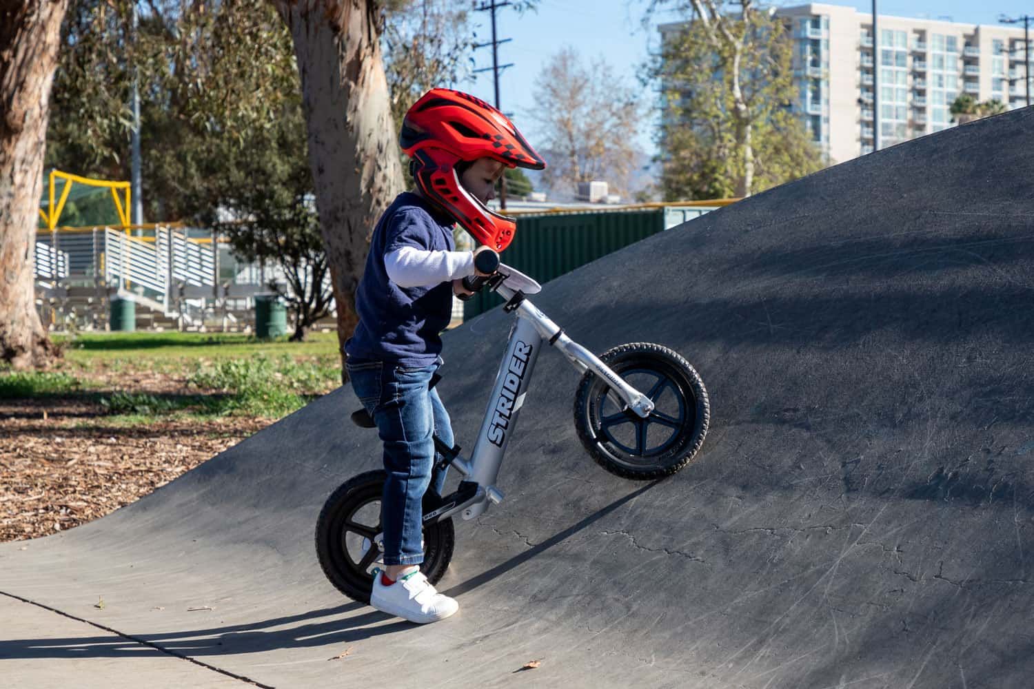 Child in a red full-face Strider ST-R helmet maneuvers a silver Strider 12 Pro balance bike on a BMX ramp