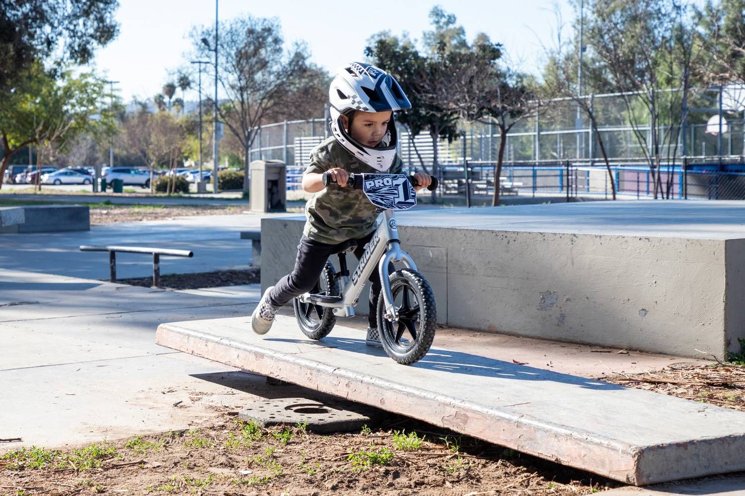 A boy in a full face bicycle helmet rides a silver Strider 12 Pro balance bike